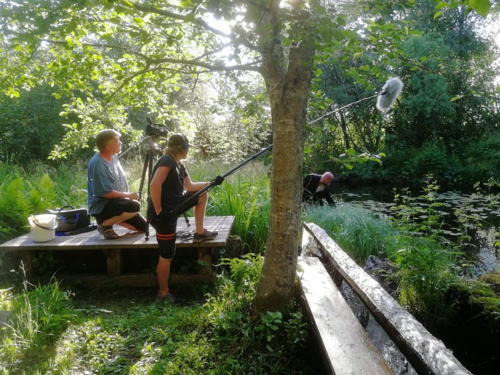 Duus-elokuvan päänäyttelijä kurkottaa joesta jotakin. On kesäisen vihreää ja aurinko paistaa. Rannalla kuvaamassa videokuvaaja ja äänimies pitelee mikkiä.